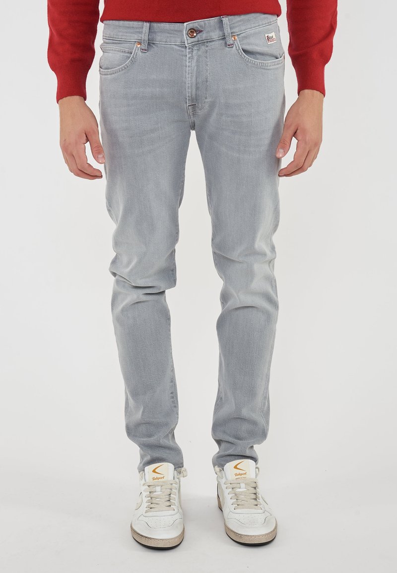 Mann mit hellgrauen Slim-Fit-Jeans, weißen Sneakers mit orangefarbenem Logo und rotem Langarmpullover, der vor einem weißen Hintergrund steht.