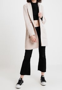 Donna che indossa un cappotto beige sopra un top nero corto, pantaloni neri a zampa d'elefante e sneakers platform nere, in piedi con una mano sul cappotto.