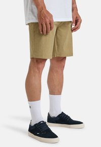 Tan shorts made from a textured fabric, knee-length with side pockets, paired with navy sneakers and white crew socks.
