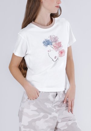 Femme portant un t-shirt blanc avec un motif floral rose et bleu et un pantalon camouflage gris clair, main dans la poche, tournée vers la droite.