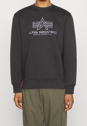 Osoba nosząca czarną bluzę Alpha Industries z logo i napisem „Knoxville, Tennessee, U.S.A.” w połączeniu z oliwkowymi bojówkami.
