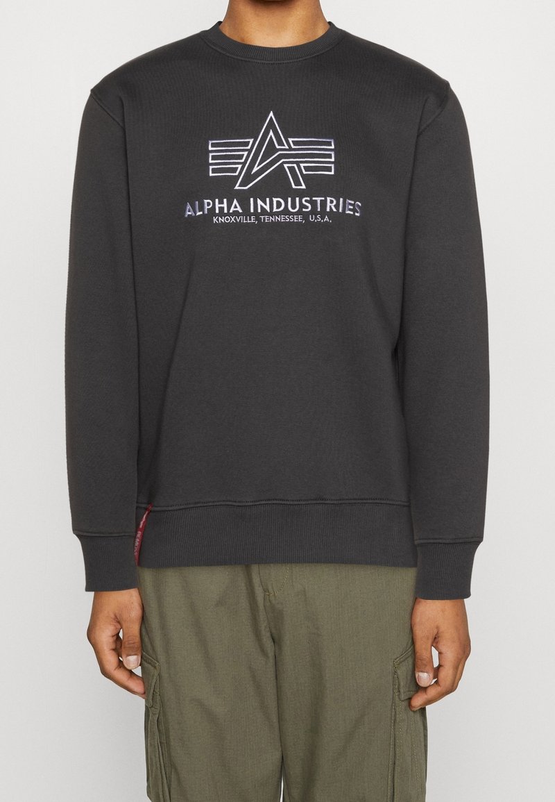 Personne portant un sweat-shirt noir Alpha Industries avec un logo et le texte « Knoxville, Tennessee, U.S.A » associé à un pantalon cargo vert olive.