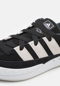 Svarta mockasneakers med vita och grå detaljer, texturerade svarta snören och en mönstrad vit gummiyttersula med triangulära detaljer.