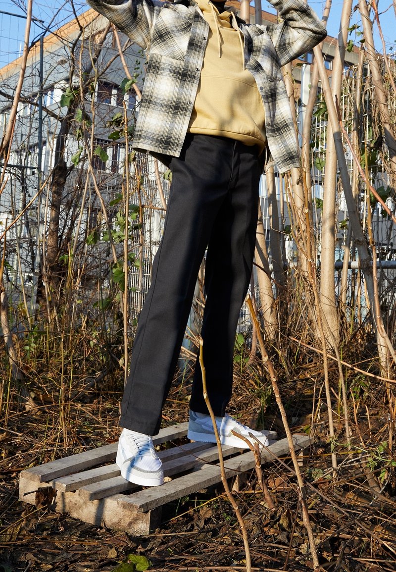 Tenue décontractée comprenant une chemise à carreaux, un hoodie beige, un pantalon noir et des baskets blanches, dans un environnement naturel et envahie par la végétation.