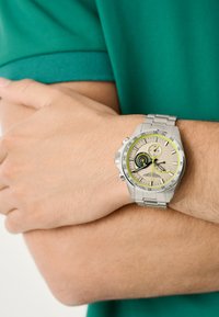 Sudraba nerūsējošā tērauda pulkstenis ar apaļu seju, bēšas ciparnīcas, melniem un dzelteniem akcentiem un metāla siksniņu, kas nēsāta uz rokas.