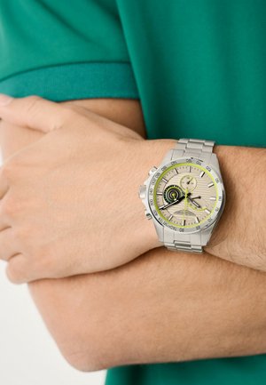 Reloj de acero inoxidable plateado con una esfera redonda, esfera beige, acentos en negro y amarillo, y una correa de metal, llevado en una muñeca.