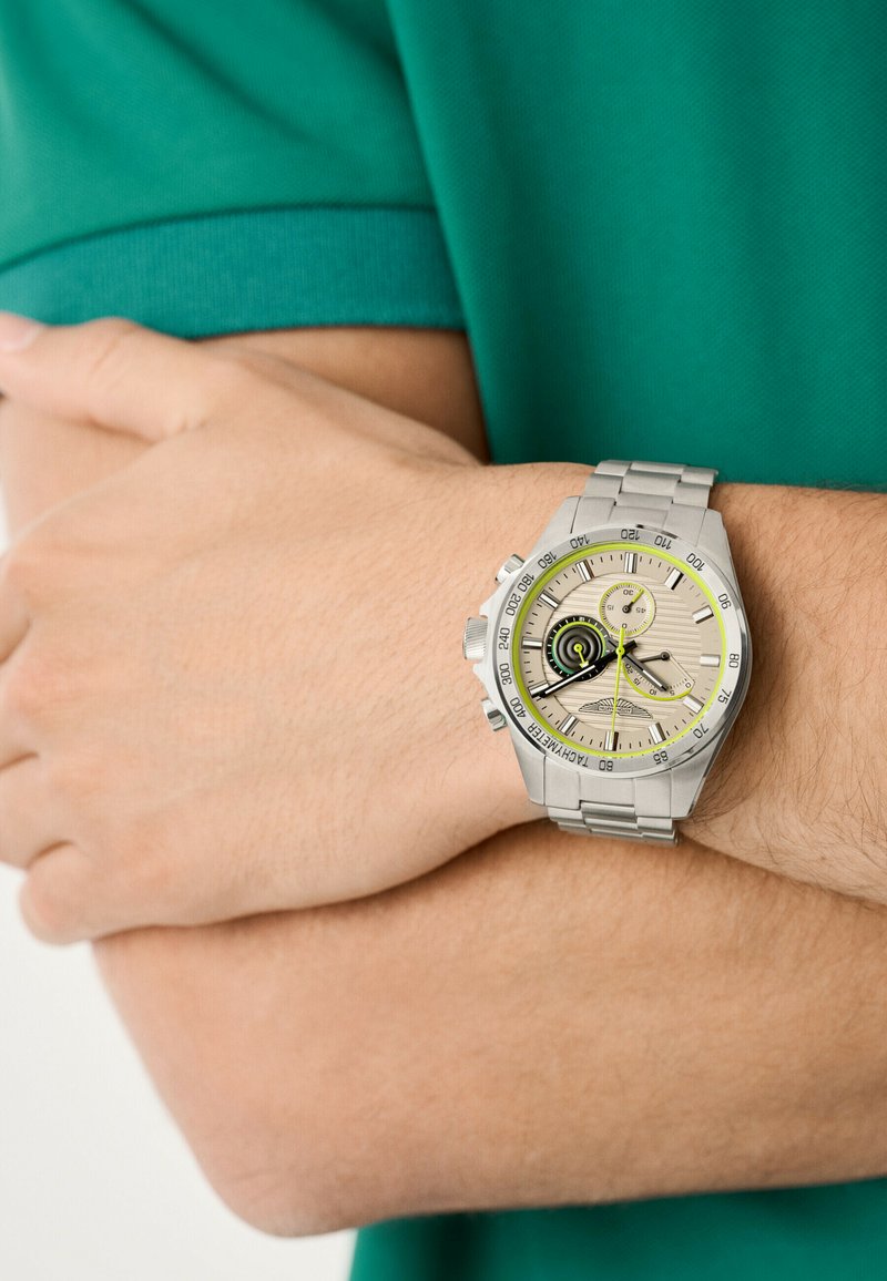 Sudraba nerūsējošā tērauda pulkstenis ar apaļu seju, bēšas ciparnīcas, melniem un dzelteniem akcentiem un metāla siksniņu, kas nēsāta uz rokas.