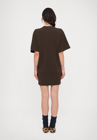 Γυναίκα που στέκεται πλάτη, φοράει ένα κοντό καφέ φόρεμα, σκούρες μπλε καλτσοδέτες και παπούτσια με λεοπάρ σχέδιο σε λευκό φόντο.