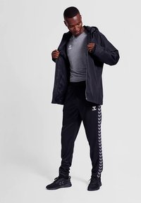 Zwarte lichtgewicht jas met een capuchon, grijs t-shirt en zwarte trainingsbroek met witte chevronstrepen en merklogo's op de borst en heup.