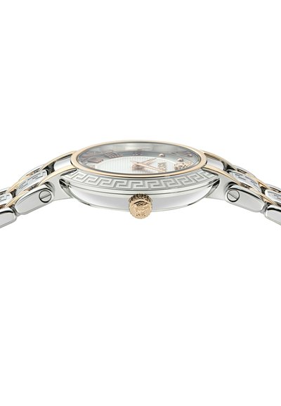 Montre argentée et dorée avec un cadran à motif, un boîtier rond et un bracelet décoratif présentant un design de cl é grecque et une couronne en forme de tête de lion.