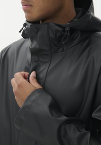 Fekete vízálló dzseki, cipzárral és patentos záródással, magas gallérral és állítható húzózsinóros kapucnival, sima, matt textúrával.