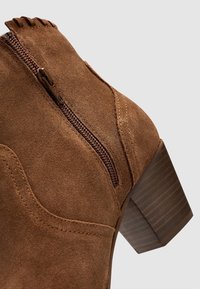 Brown suede ankle boot with a decorative zipper and a stacked heel. Features curved stitching and a textured surface.