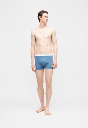 Mladý muž stojaci naboso, oblečený v modrých boxerkách Calvin Klein, pozerajúc sa doľava na jednoduchom svetlom pozadí.