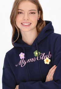 Sudadera con capucha azul marino con flores bordadas en púrpura, verde, amarillo y naranja. Presenta "MymoClub" en letras cursivas rosas.