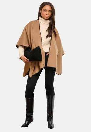 Beiger Wollponcho mit schwarzen genähten Kanten, über einem cremefarbenen Rollkragenpullover getragen. Dazu eine schwarze, figurbetonte Leggings und kniehohe Stiefel. Schwarze Clutch.