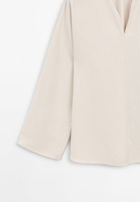 Blouse beige clair avec un décolleté en V, des manches longues et larges, et un tissu légèrement texturé. Présente un ourlet droit et des fentes sur les côtés pour faciliter le mouvement.