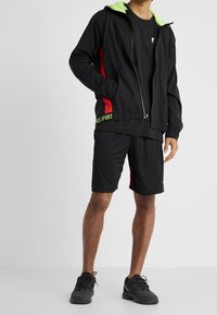 Mies wearing a black sports jacket with a green hood lining, black shorts with red side panels, a black t-shirt, and black athletic shoes.