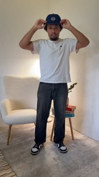 T-shirt branca, jeans escuros de ganga, ténis preto e branco, boné azul-marinho com detalhe bordado. Em pé numa sala com textura suave perto de uma cadeira.