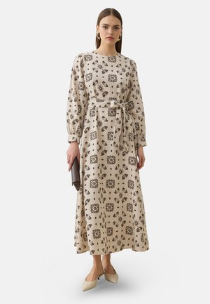 Kvinde iført en beige langærmet blomsterkjole med et bundet bælte, holder en mørkebrun clutch og har beige spidse sko på.