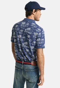 Marineblauw poloshirt met wit floral en script patroon; korte mouwen en kraag; gecombineerd met vervaagde blauwe jeans en een donker pet.