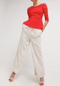 Top peplum en dentelle rouge avec un motif texturé, associé à un pantalon large blanc. Le modèle porte des sandales à talons nudes.