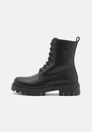 Black leather ankle boot with a round toe, lace-up closure, textured pattern, and a thick rubber sole. Features logo detail on the side.