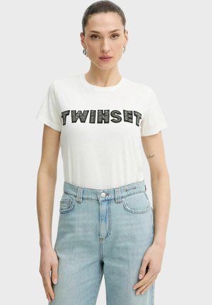 Donna con capelli raccolti indossa una maglietta bianca con la scritta nera "TWINSET" e jeans a vita alta azzurri chiaro, su uno sfondo semplice.