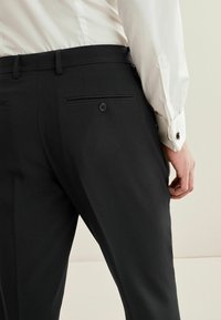 Schwarze Hosen aus glattem Stoff, schlank geschnitten, mit zwei hinteren Taschen mit Knöpfen und flacher Frontgestaltung. Hemd mit sichtbarem Knopfverschluss.
