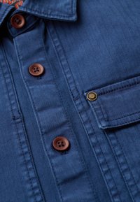 Camicia blu a motivo a spina di pesce con bottoni marroni, tasca con pattina e un tessuto texture. Dettagli di cucitura notevoli sono visibili.