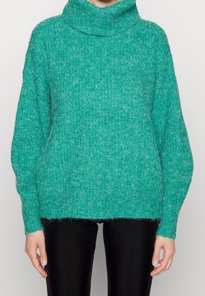 Personne portant un pull à col roulé en maille épaisse verte et un pantalon noir, debout devant un fond clair uni.