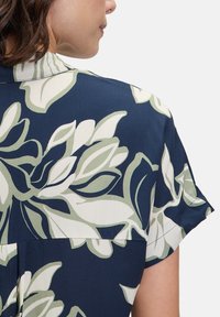 Blumenbluse mit navy Hintergrund, die große grüne und weiße Blumenmuster zeigt. Kurze Ärmel und einen kragenförmigen Ausschnitt. Glatte Textur.