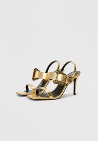 Gold metallic high-heeled sandals featuring a square toe, thin straps, and a decorative buckle with chain detail. Smooth texture, elegant design.