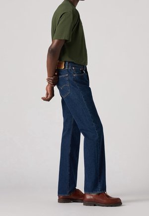 Personne portant un jean bleu foncé droit, une chemise à manches courtes vert olive et des chaussures en cuir marron, debout de côté avec les mains dans le dos.