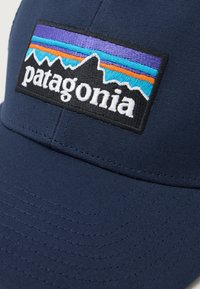 Boné azul-marinho feito de tecido com um design estruturado. Apresenta um retângulo preto com um logótipo de montanha multicolorido e o texto "patagonia".