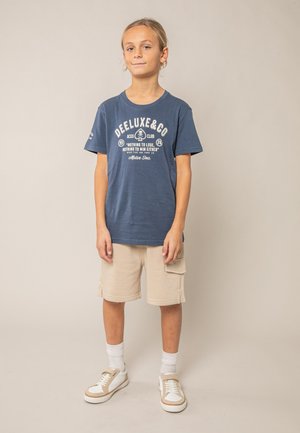 Kind met lang haar, gekleed in een blauw T-shirt met opdruk, beige cargoshorts, witte sokken en beige-witte sneakers, staand tegen een effen achtergrond.