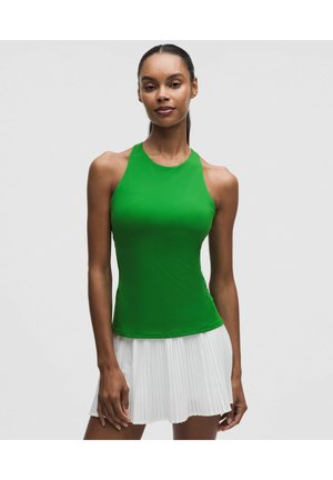 Femme portant un haut sans manches vert vif et une jupe plissée blanche, debout sur un fond gris clair uni.