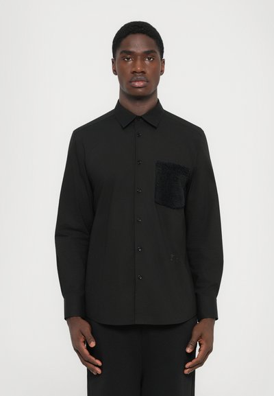 Chemise noire à manches longues avec un col rabattable, dotée d'une poche à rabat en fausse fourrure noire texturée sur la poitrine gauche. Tissu lisse et léger.