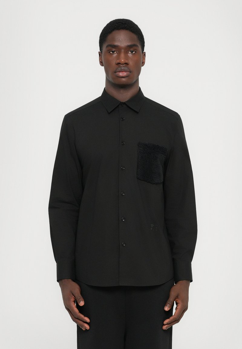 Chemise noire à manches longues avec un col rabattable, dotée d'une poche à rabat en fausse fourrure noire texturée sur la poitrine gauche. Tissu lisse et léger.