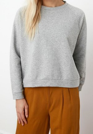 Vrouw die een lichtgrijze longsleeve trui en losse, hooggetailleerde bruine broek draagt, staand voor een effen witte achtergrond.