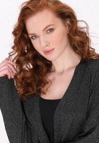 Černá, třpytivá šála s volnými, splývavými rukávy a hlubokým výstřihem do V. Modelka má vlnité červené vlasy a pod tím jednoduchý černý top.