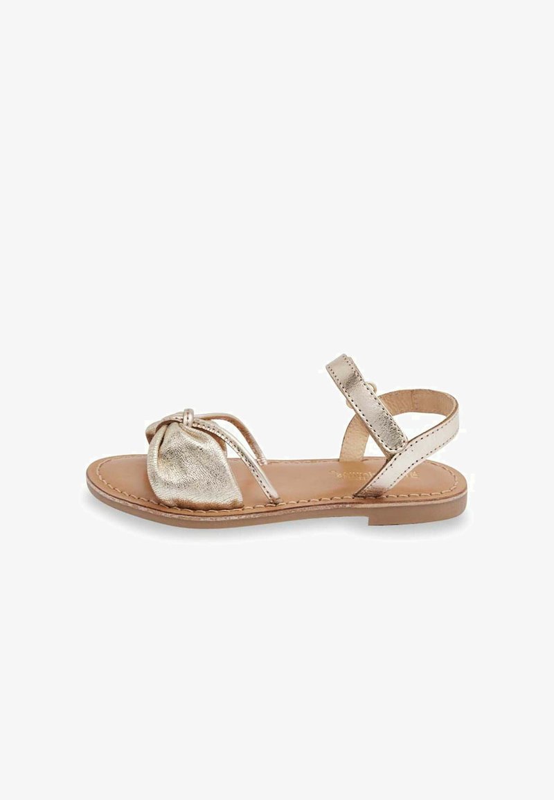 Sandal en métal doré avec un design à l'avant noué. Dispose d'une semelle plate, d'une bride de cheville et de découpes sur les côtés.