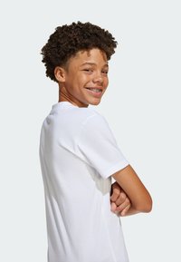 Weißes Baumwoll-T-Shirt mit rundem Halsausschnitt und kurzen Ärmeln. Das Model hat eine lockige Frisur und steht seitlich mit verschränkten Armen.