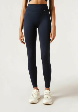 Marineblaue Leggings aus dehnbarem Stoff, mit hohem Bund und einem kleinen Logo-Detail, kombiniert mit weißen Sportschuhen.