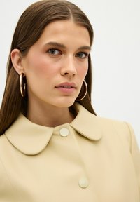 Beige Mantel mit Kragen und zwei runden Knöpfen, der eine glatte Textur aufweist. Das Model trägt große goldene Creolen.