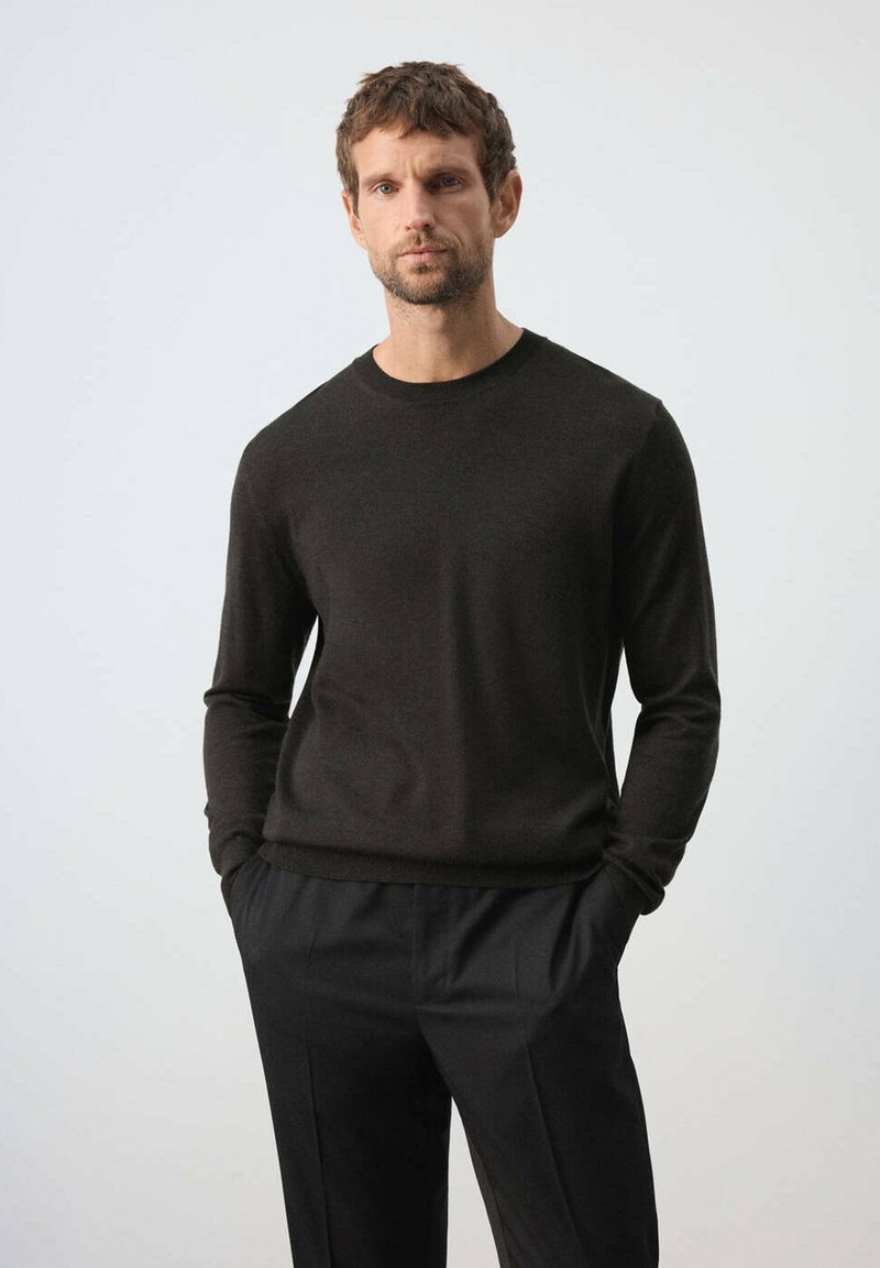 Homme debout, les mains dans les poches, portant un pull noir à manches longues et un pantalon noir sur un fond clair uni.