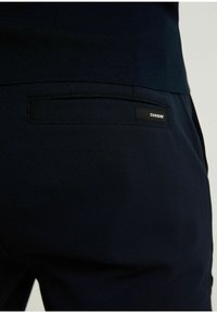 Donkerblauwe broek met een gestructureerde afwerking, voorzien van een achterzak en een klein zwart label met de tekst "CHASIN'".