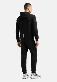 Sudadera con capucha negra y pantalones jogger a juego, con un diseño minimalista y rayas horizontales blancas cerca de la cintura, fabricados en un tejido suave.
