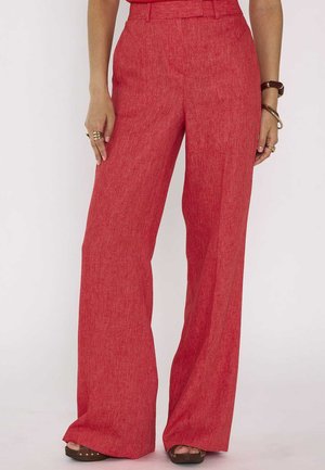 Femme portant un pantalon rouge taille haute à jambes larges associé à des sandales ouvertes marron, montrant le bas du torse jusqu'aux pieds et les mains sur les côtés.