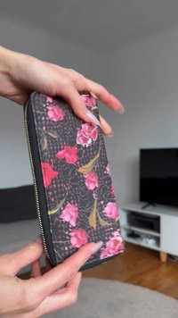 Floral wallet featuring a black background, pink flowers, and green leaves. Solid black zipper and textured fabric. Rectangular shape.