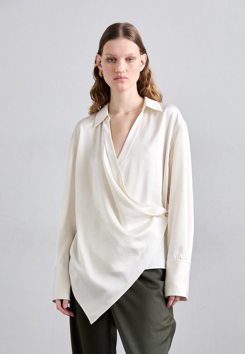 Blusa branca assimétrica com um decote em V profundo, mangas longas e frente drapeada. Feita de tecido liso, apresenta um detalhe em botão de um lado.